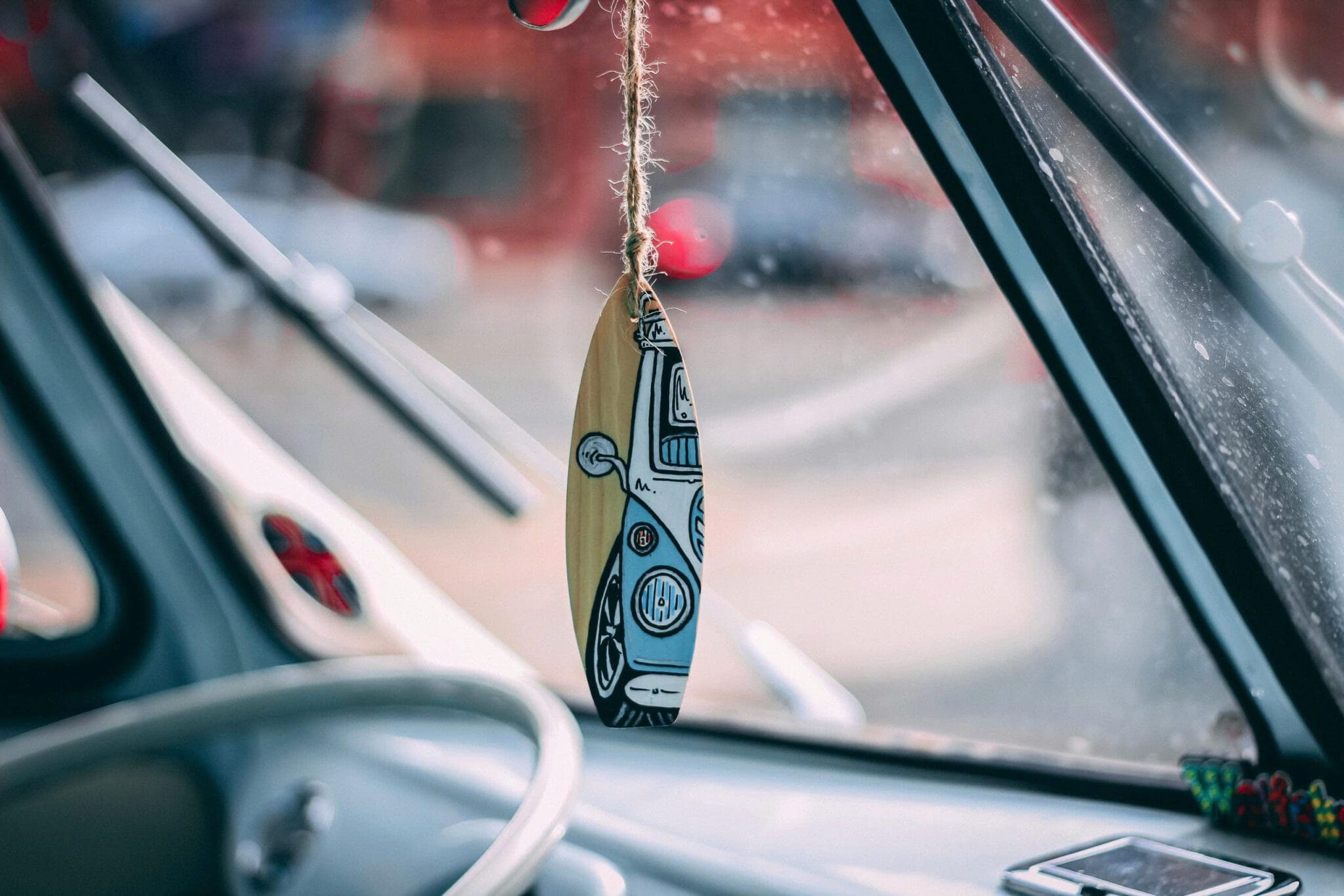 Wooden surfboard shaped ornament illustrated with a camper van, hanging from the mirror inside a vintage van looking out through the windshield