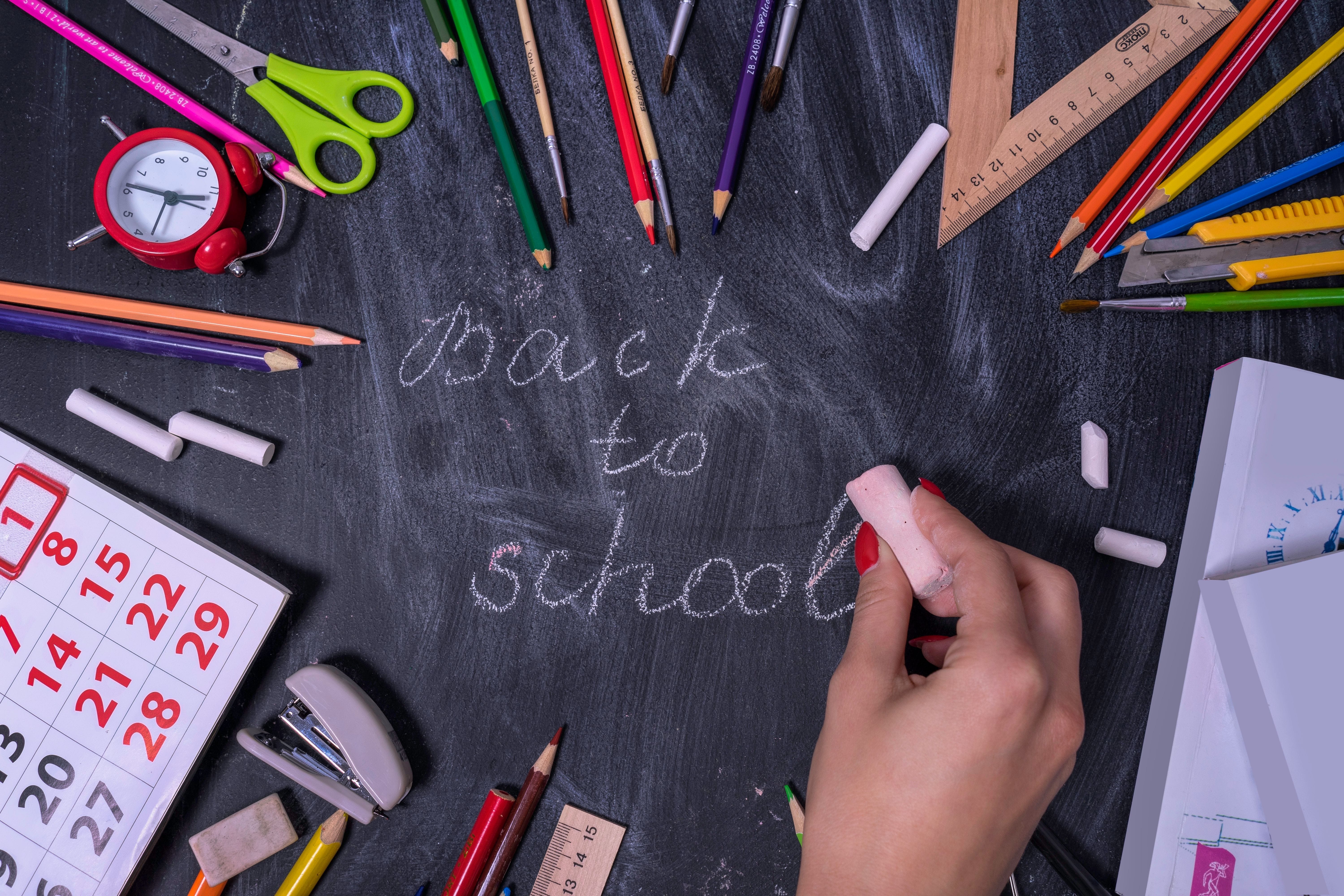 Back to School written in chalk on a blackboard surrounded by pencils, scissors, chalk, a ruler and a red alarm clock