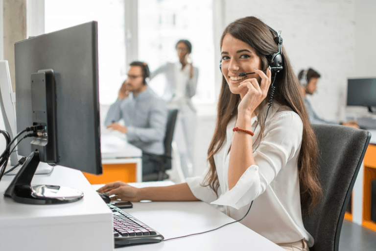 Smiling customer service agent wearing a headset at a desk with a monitor and keyboard, colleagues working behind her