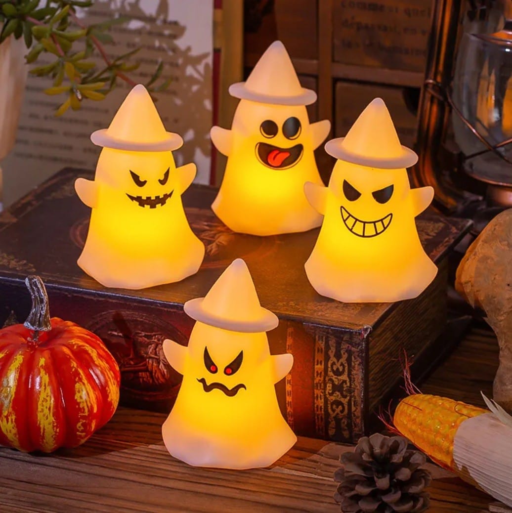 Four battery-powered LED ghost lights in witch hats glowing on a wooden table beside a pumpkin
