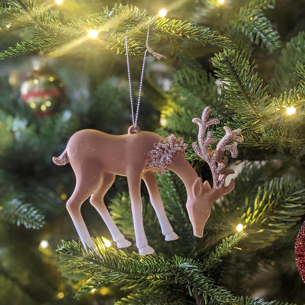 Pastel deer ornament hanging from a fir branch beside gold baubles and warm string lights