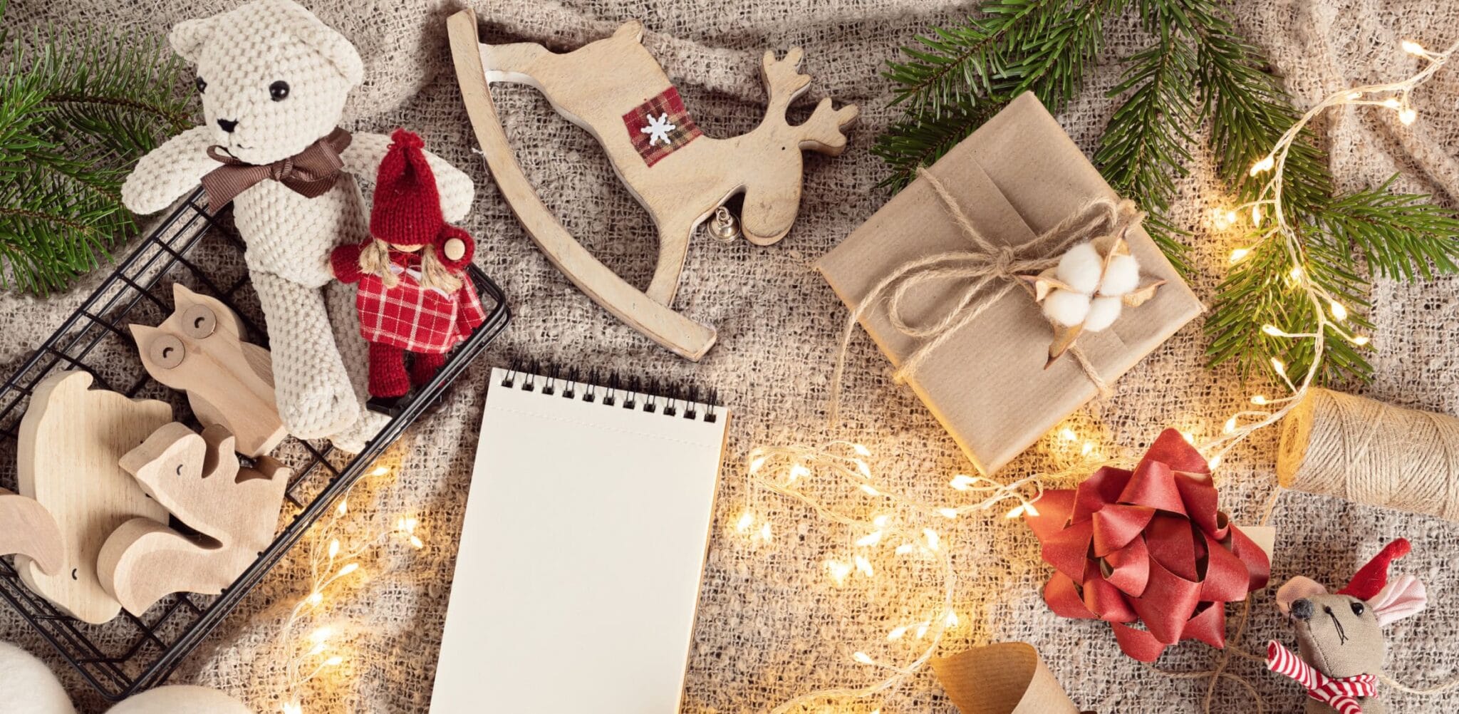 Flat lay on burlap with a knitted teddy bear, wooden animal figures, a kraft paper gift box, a blank notepad and string lights
