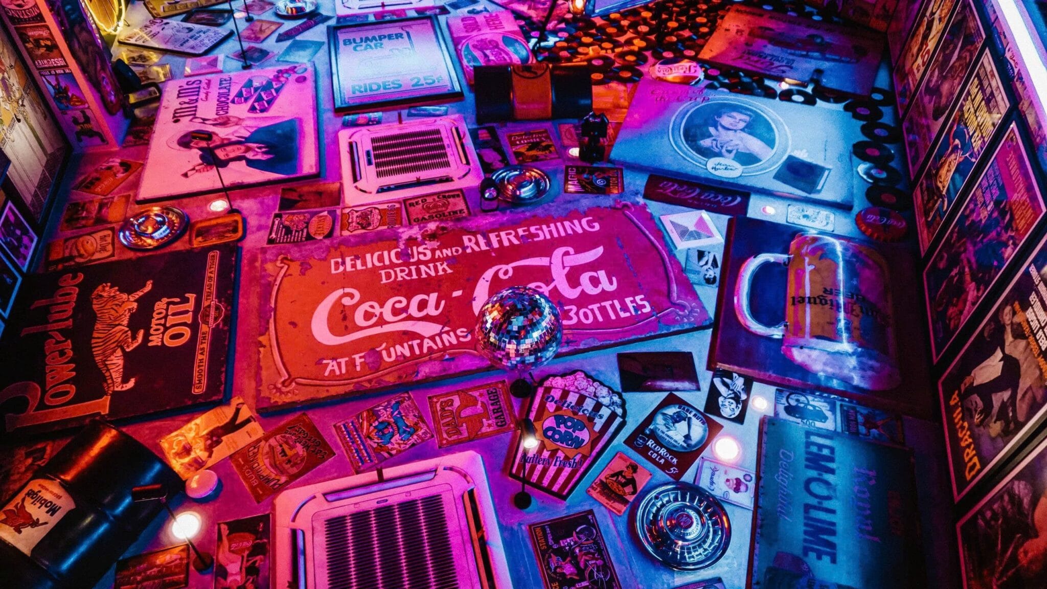 Ceiling and walls covered in vintage advertising signs, posters, vinyl records and a disco ball under pink and purple neon light