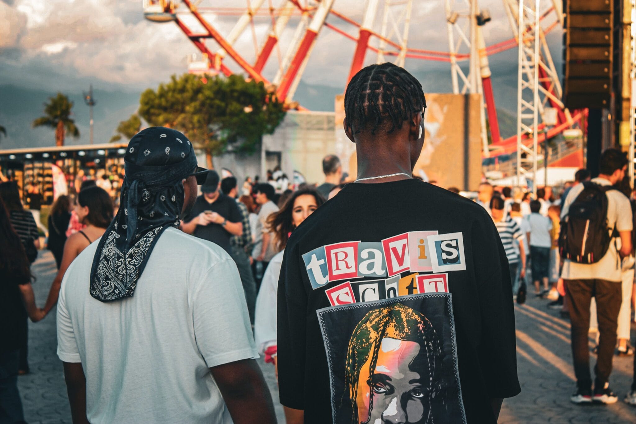 Two people seen from behind walking through a crowded fairground at sunset, one wearing an oversized graphic t-shirt, ferris wheel in the background