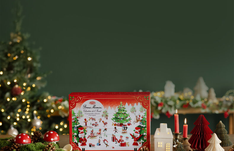 Red illustrated Advent calendar box standing on a wooden table among toadstool ornaments, pinecones, candles and paper trees