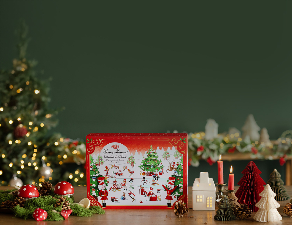 Red illustrated Advent calendar box standing on a wooden table among toadstool ornaments, pinecones, candles and paper trees