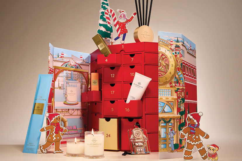 Red beauty Advent calendar with numbered pull-out drawers, opened to reveal a lit candle, a body cream tube and a reed diffuser, framed by gingerbread cutouts